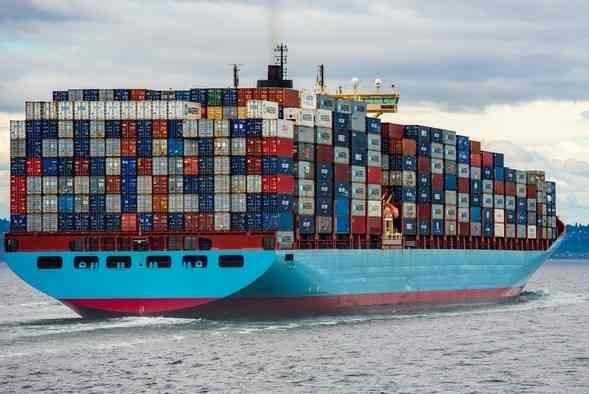 A ship piled high with shipping containers sails out of a port