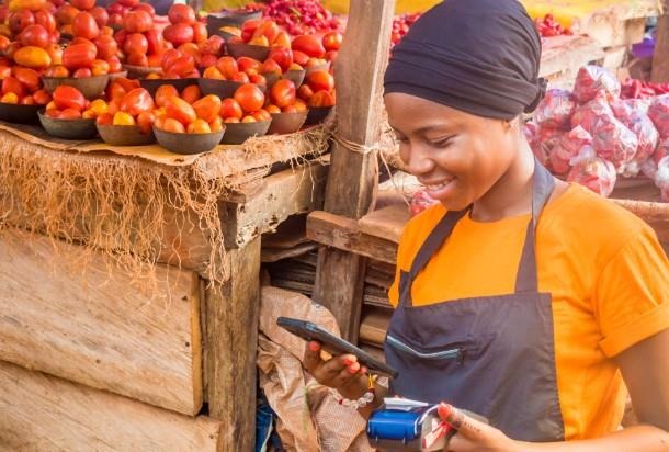 Young African woman selling in a local market holding a mobile point of sale system and using her mobile phone