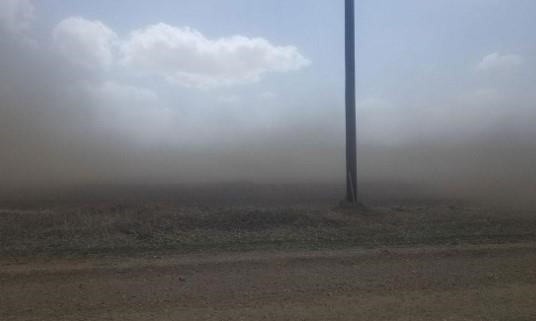 Blue sky with some clouds. Lower horizon is a tilled field with dirt/dust in the air beneath the clouds, actually blackening out the blue sky near the earth’s surface